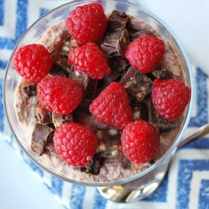 overhead photo of a chocolate ricotta cream base topped with a chopped protein bar and fresh raspberries in a parfait glass over a blue Chevron napkin