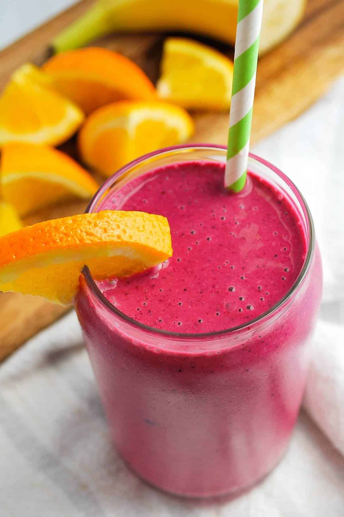 overhead shot of a banana beet smoothie with an orange slice in a glass with a straw with sliced orange and banana behind it