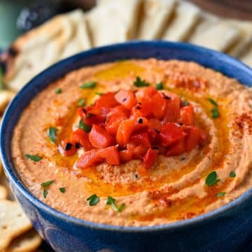 close up of a bowl of paprika roasted red pepper hummus with evoo and parsley on top with chopped red peppers and pita slices behind it