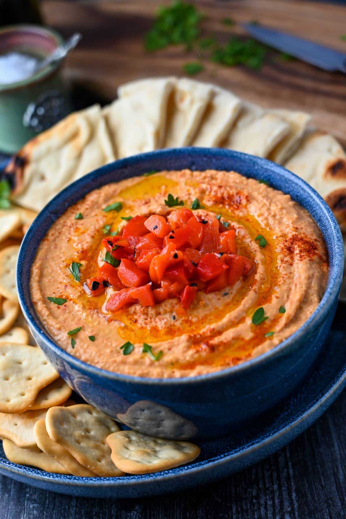 paprika hummus in a blue serving bowl topped with roasted red peppers and served with crackers and pita bread