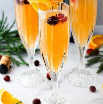 four prosecco mimosas in champagne glasses with fresh cranberries floating on top and an orange slice on the rim