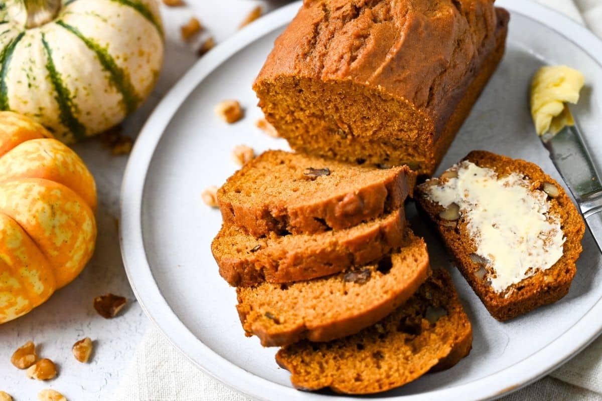 a sliced pumpkin bread loaf with a piece with butter on it next to a knife with mini pumpkins in the background