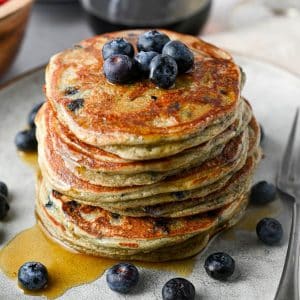 close up of a tall stack of blueberry pancakes with blueberries and syrup around it and on top