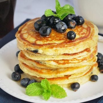 blueberry pancakes stacked on a white plate topped with fresh blueberries, syrup, and fresh mint sprigs
