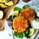 two salmon patties on a white plate with lemon wedges and mixed greens