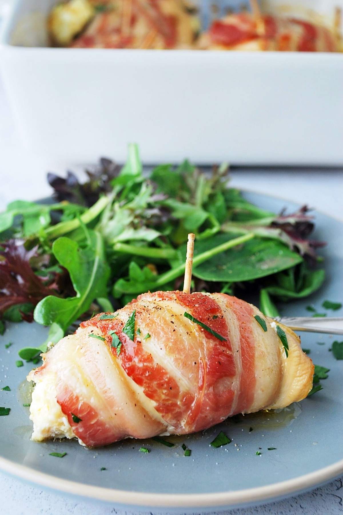 a bacon-wrapped chicken breast on a plate with a side salad and a pan of bacon-wrapped chicken breasts in the background