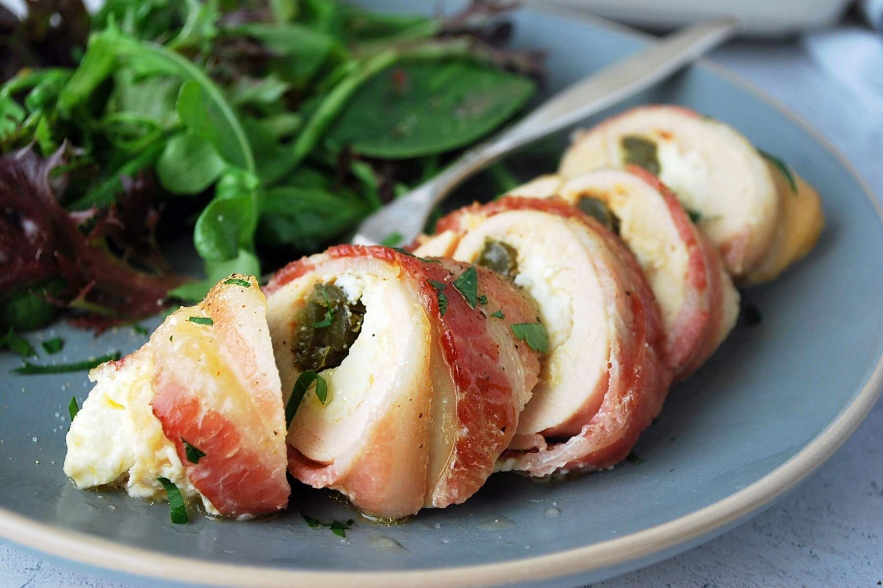 sliced stuffed chicken breast wrapped in bacon on a plate with salad and a fork