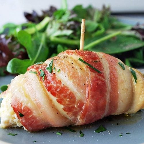 close up of a bacon-wrapped chicken breast sprinkled with fresh parsley on a plate with greens