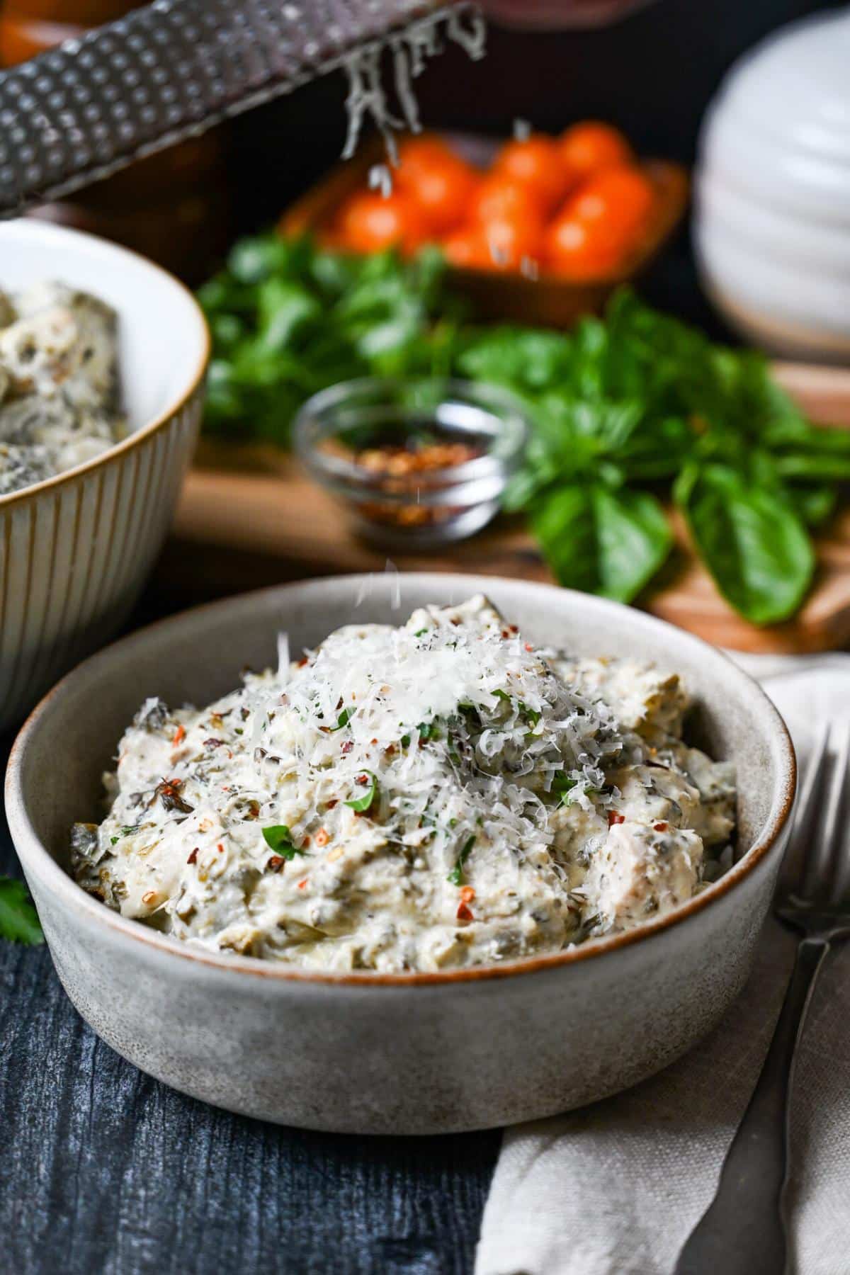 bowl of spinach artichoke chicken with fresh parmesan and basil on top with tomatoes and basil in the background