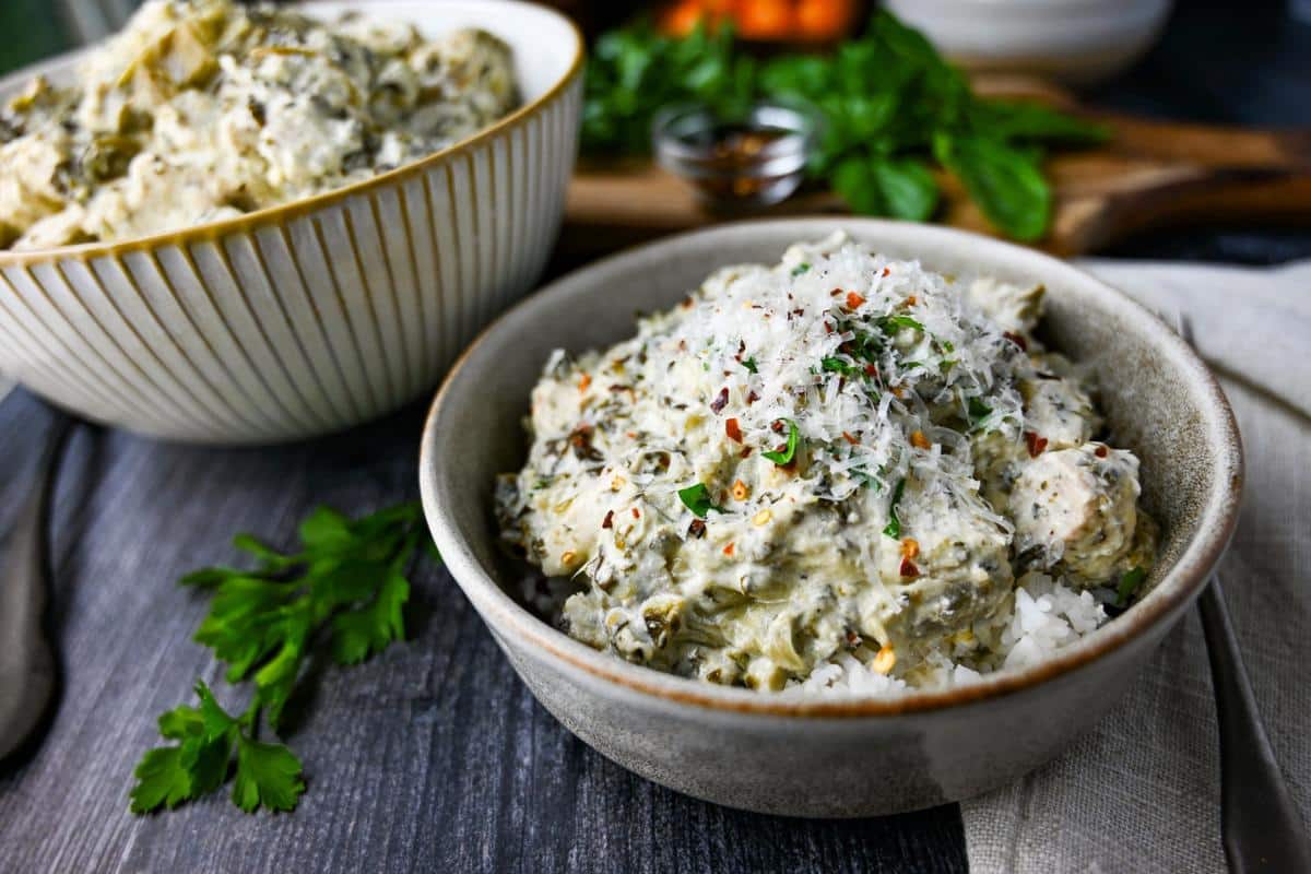 a served bowl of creamy artichoke spinach chicken with a bowl of it in the background