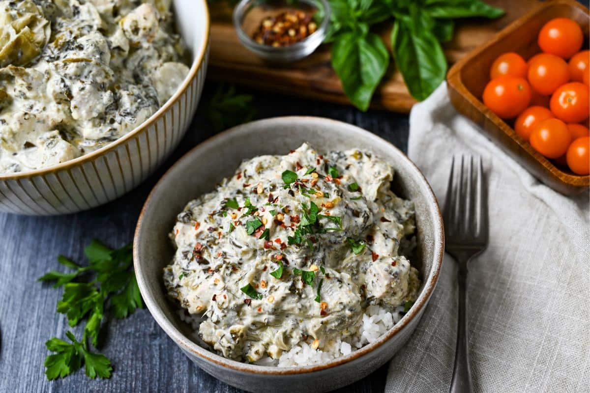 a serving bowl of creamy chicken with spinach and artichoke hearts with a served bowl with a napkin and fork