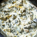 Close up of cooked chicken with spinach and artichokes in a slow cooker with a serving spoon