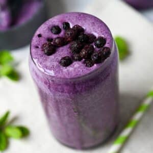 overhead photo of a blueberry protein shake with frozen blueberries and sprigs of mint around the glass