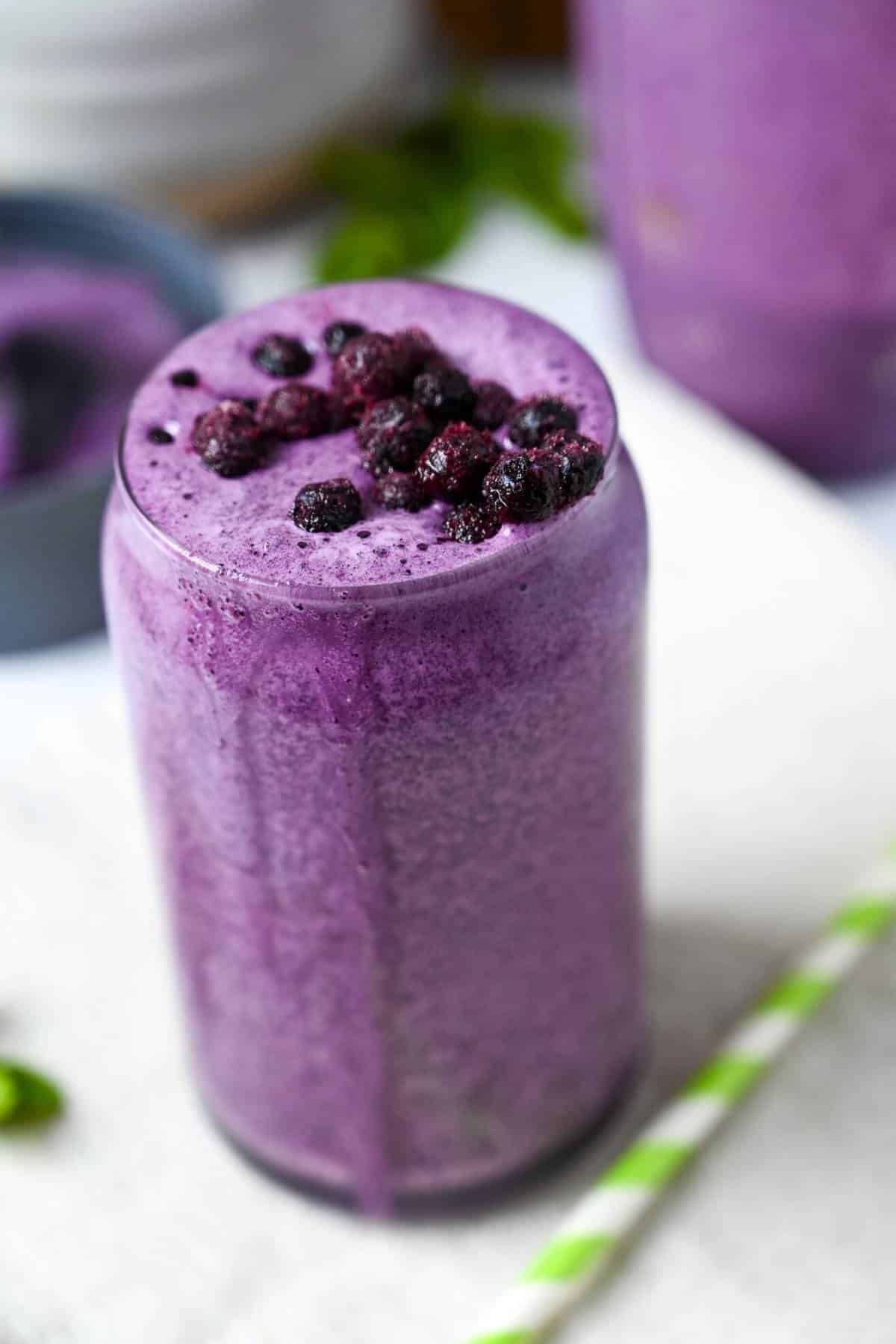 a blueberry protein shake in a glass topped with frozen wild blueberries with a straw next to it