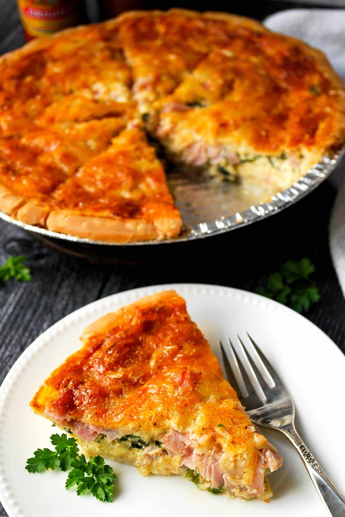 A slice of quiche plated with the whole quiche behind it