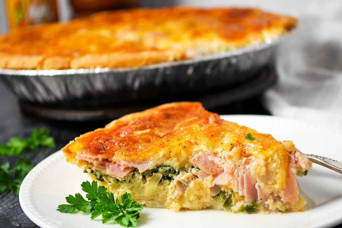 a side view of a slice of quiche with a pan of quiche in the background
