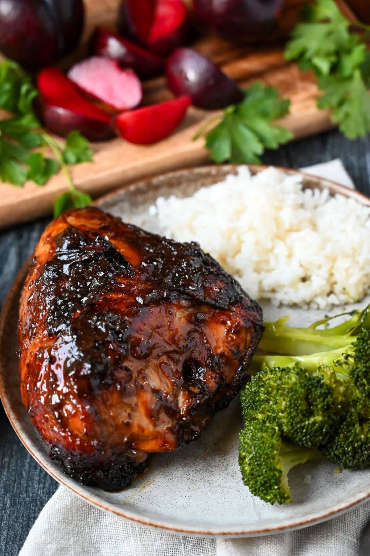 a grilled BBQ chicken breast on a plate with rice and a green veggie with plums and parlsey on a cutting board