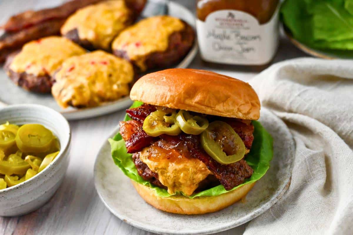 a burger on a plate with bacon, jalapenos, maple bacon jam, and lettuce, with a bowl of jalapenos and plate of burgers