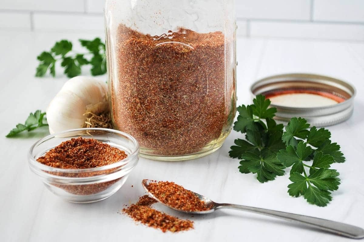 a jar of taco seasoning with a lid with a clove of garlic and fresh parsley and a dish and spoon around it