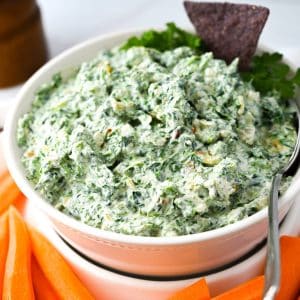close up of a bowl of spinach dip with carrots around it and a blue corn chip sticking in the top