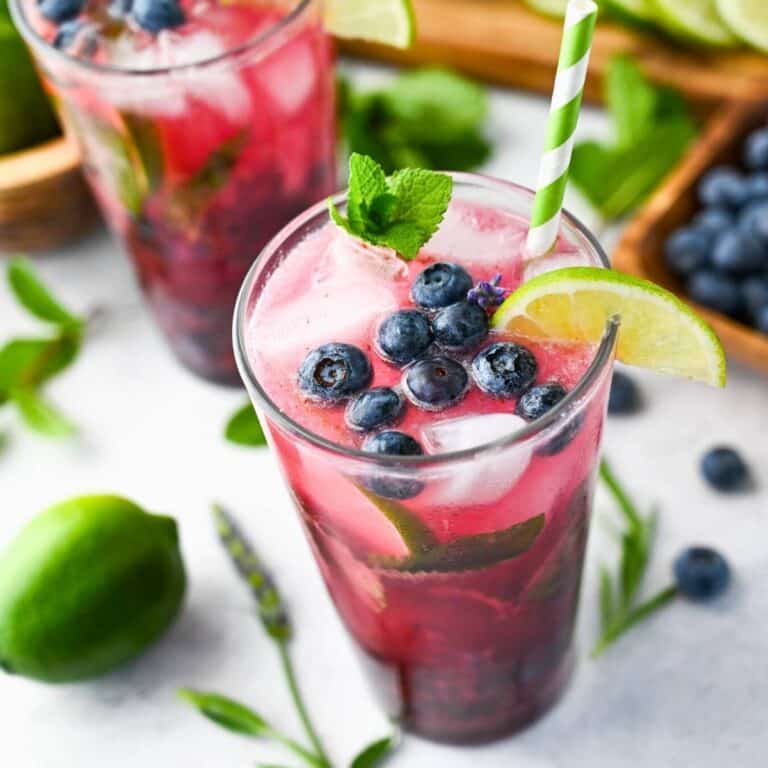 blueberry lavendar mojitos in glasses with a straw with sprigs of lavendar fresh blueberries, and limes around them