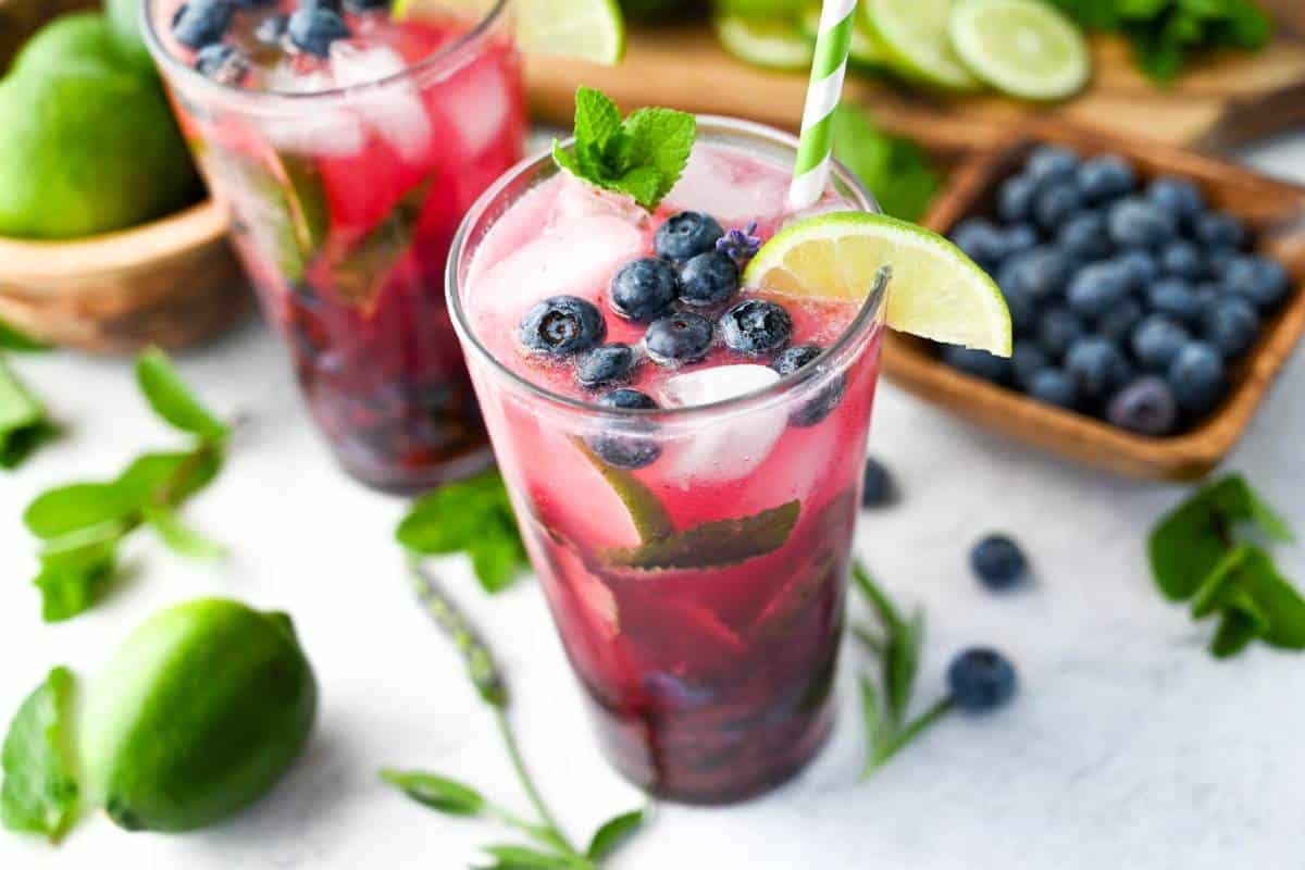a bowl of blueberries next to cocktails with fresh lavender and limes around it
