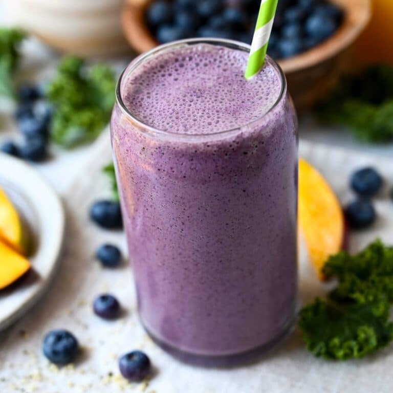 a wild blueberry smoothie made with kale, oj, and mango in a glass with blueberries, kale, and mango around it