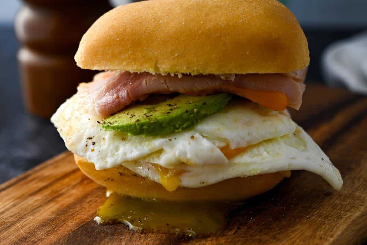 a gluten free sandwich with eggs, cheese, ham, avocado, and drippy yolk on a wood board
