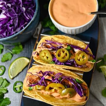 two fish tacos topped with red cabbage, jalapenos and chipotle sauce