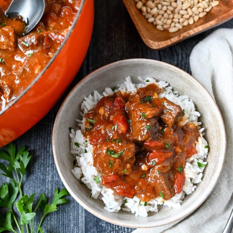 overhead view of a bowl of mango beef curry over rice with a dutch oven full of mango curry beef stew and a bowl of pine nuts