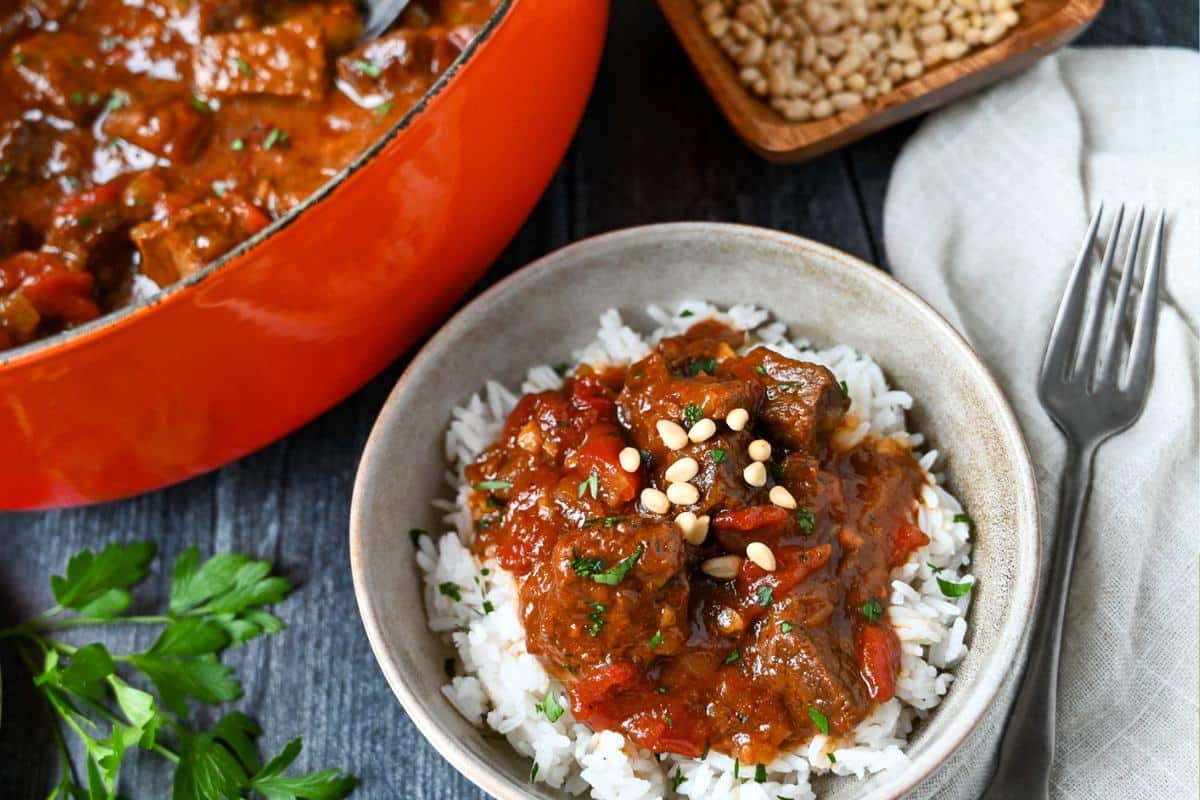 a dutch oven of stew with a bowl of stew over rice and fresh parsley and pine nuts