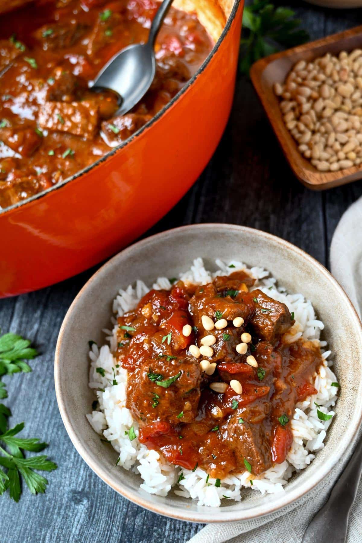 a photo of beef stew in a dutch oven with a spoon with a served bowl of mango curry beef stew over rice with parsley and pine nuts