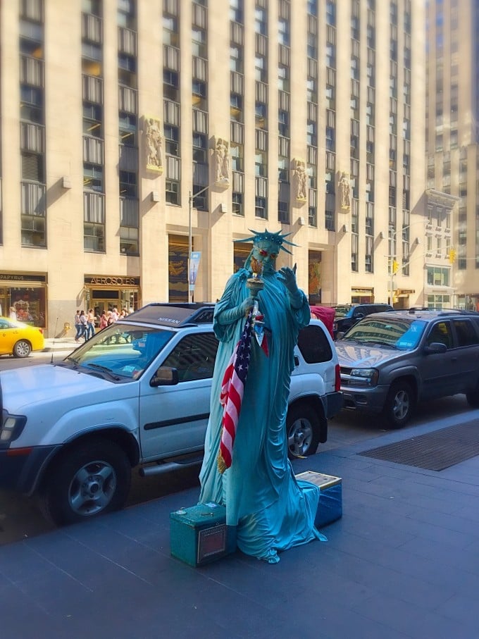 A car parked on a city street with a woman dressed up as the Statue of Liberty in front