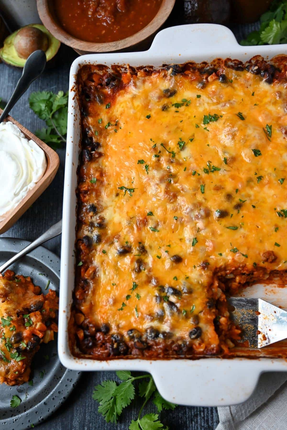 looking down at a pan of mexican casserole with a served slice on a plate and bowl of sour cream