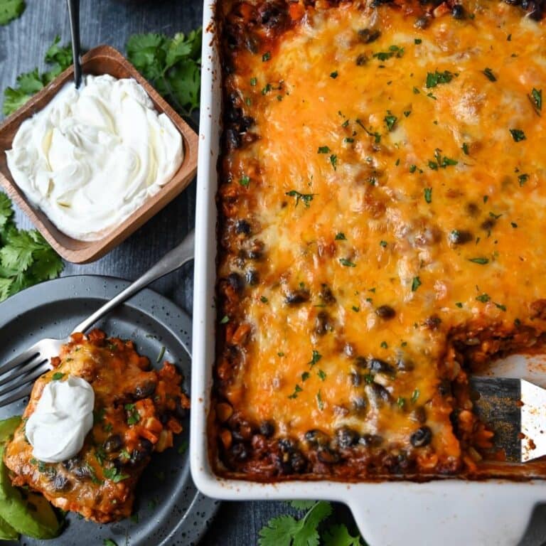 a pan of ground beef mexican casserole with a slice cut out and a spatula with a served plate of casserole topped with sour cream