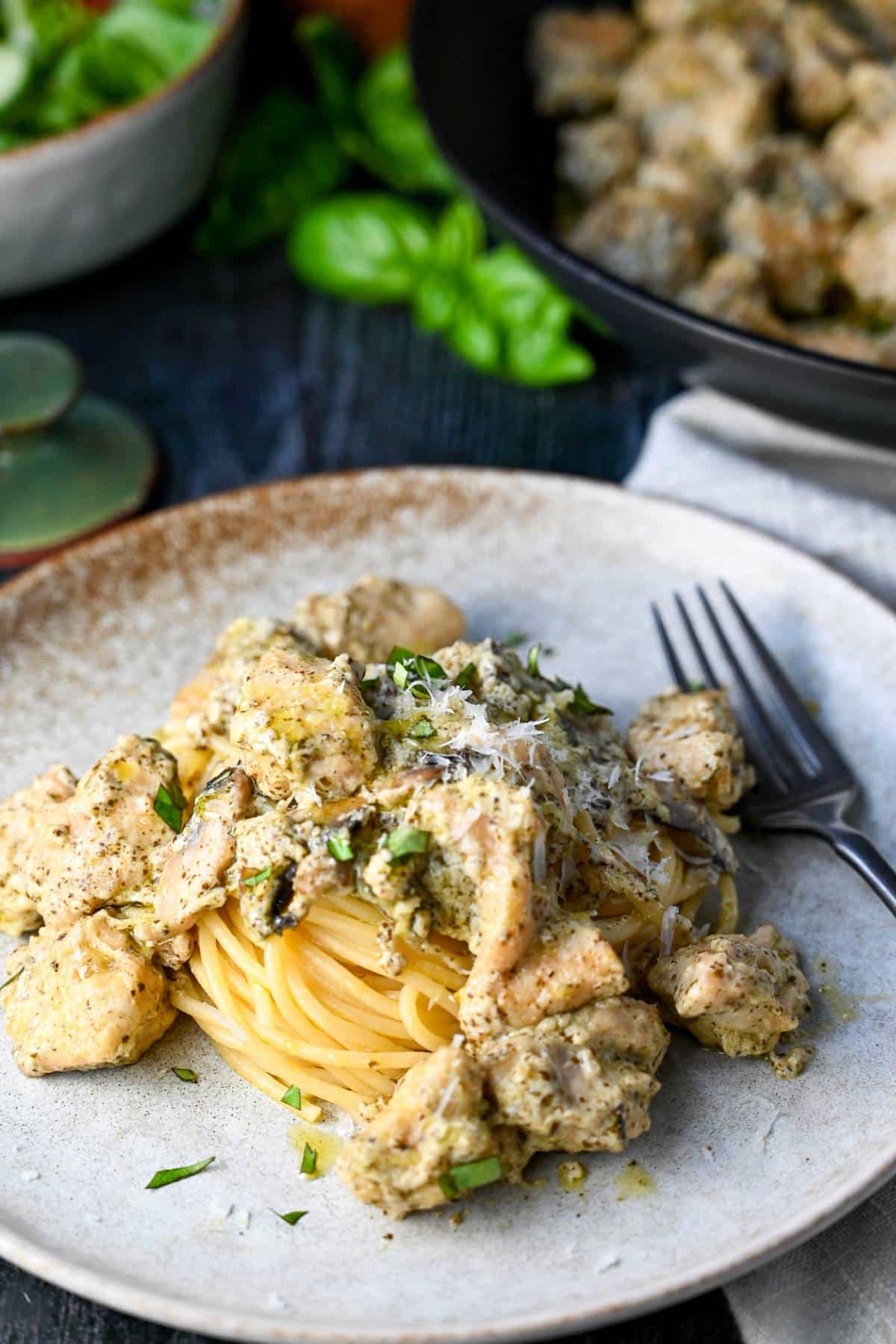 bite size chicken cooked in a pesto sauce on top of a bed of spaghetti noodles with fresh basil on top