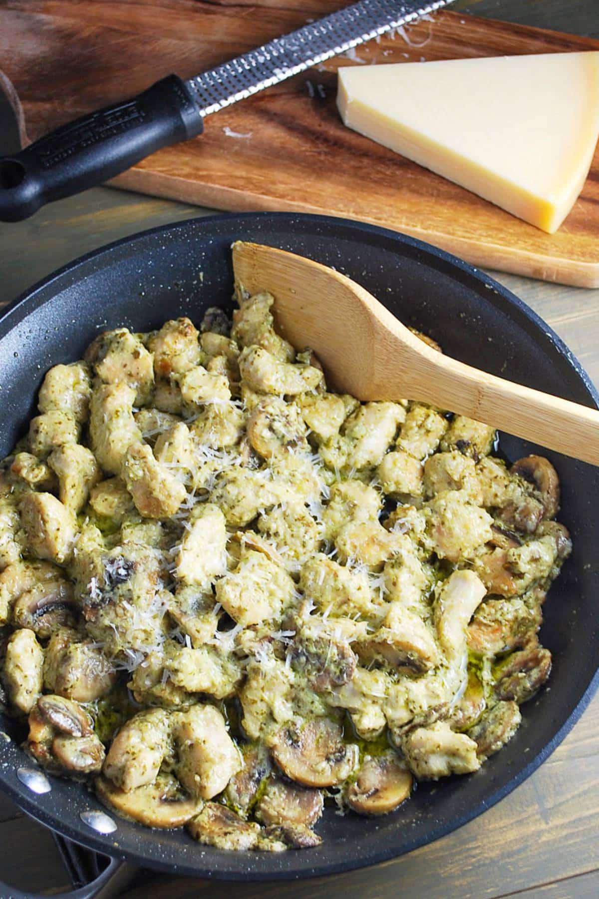 a skillet of chicken in pesto sauce with a block of parmesan cheese on a cutting board