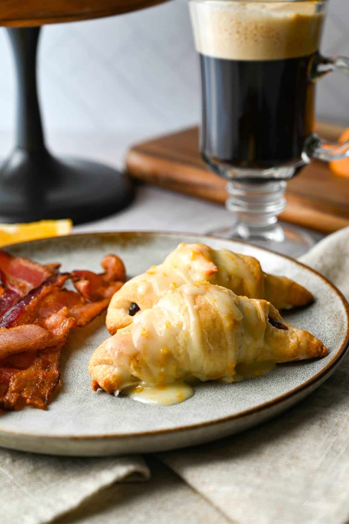 two croissants dripping with orange glaze with bacon on a plate with a cup of coffee behind it