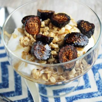 yogurt in a parfait glass topped with granola, sliced figs, and maple syrup with a blue chevron patterned napkin underneath