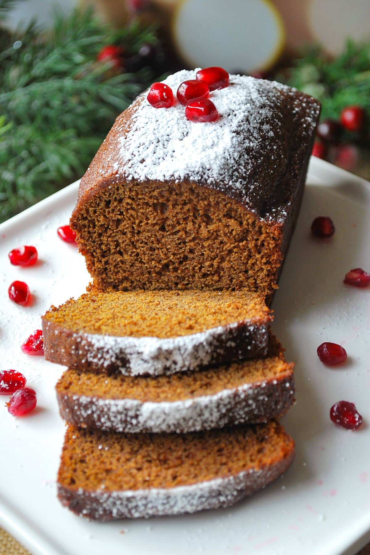 a mini loaf of gingerbread sliced on a platter topped with powdered sugar and pomegranate seeds