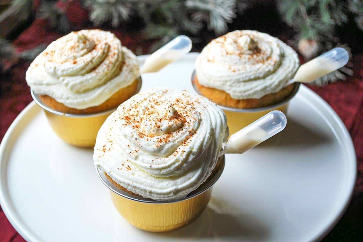 three egg nog cupcakes topped with whipped cream and grated nutmeg with greenery in the background