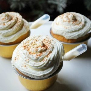 three cupcakes on a platter with pipettes of egg nog and topped with whipped cream icing and grated nutmeg