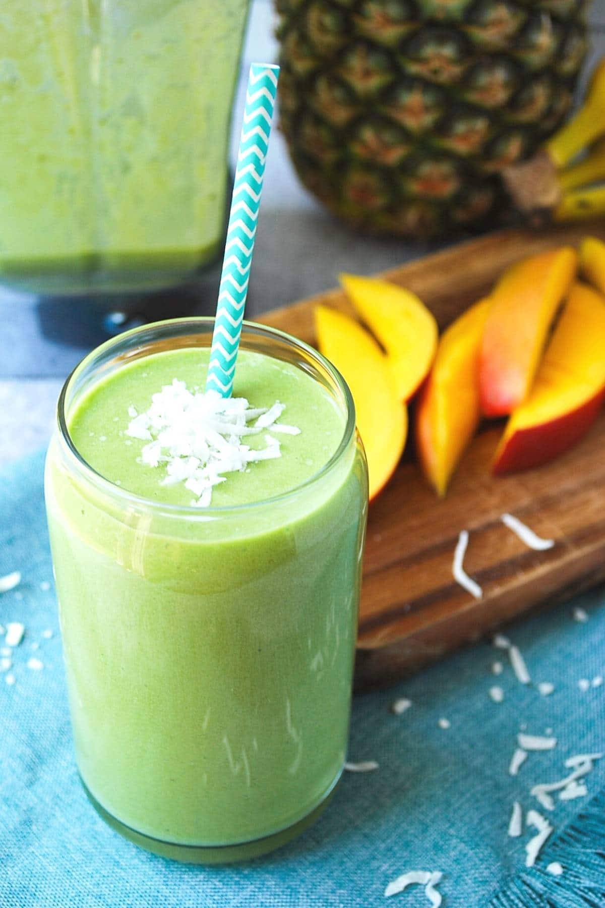 island green smoothie in a glass topped with coconut with sliced mango and a pineapple in the background