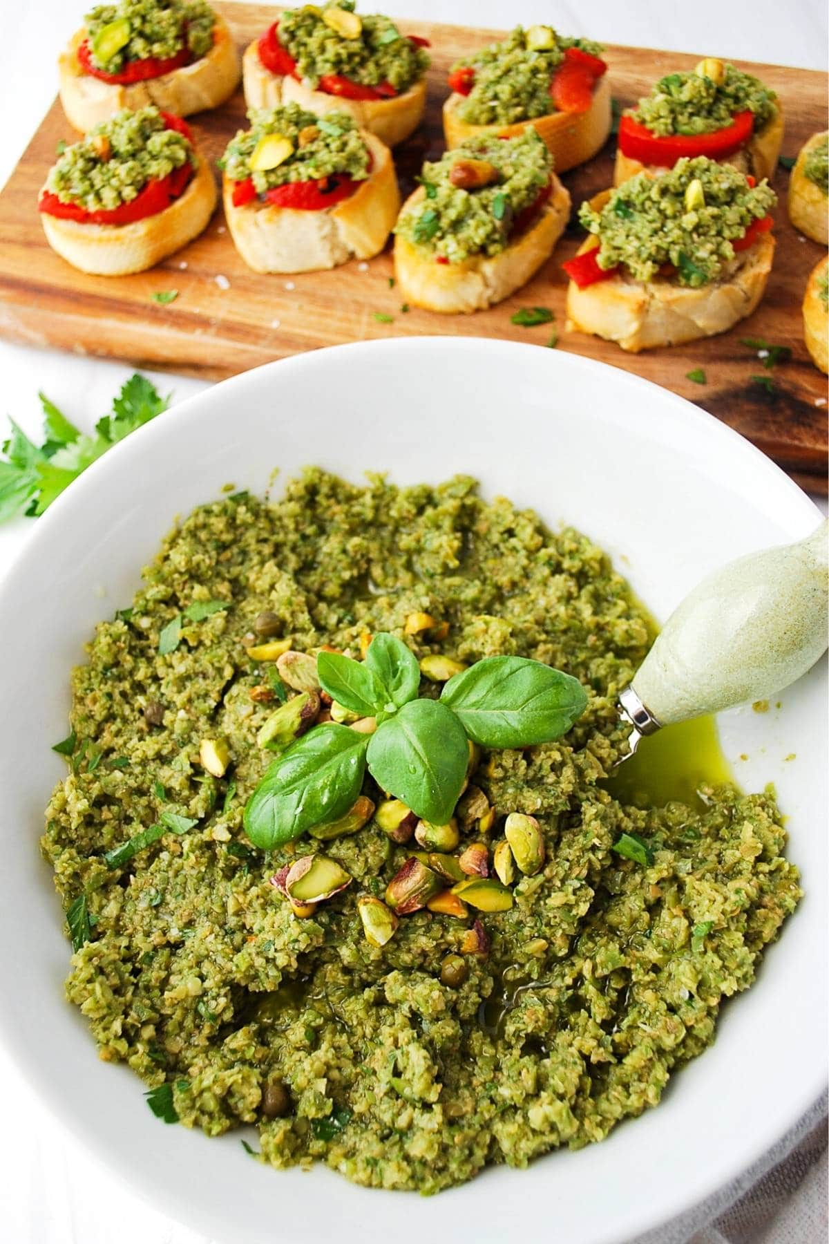 overhead photo of a serving bowl with green olive tapenade topped with basil and pistachios with brushetta behind it.