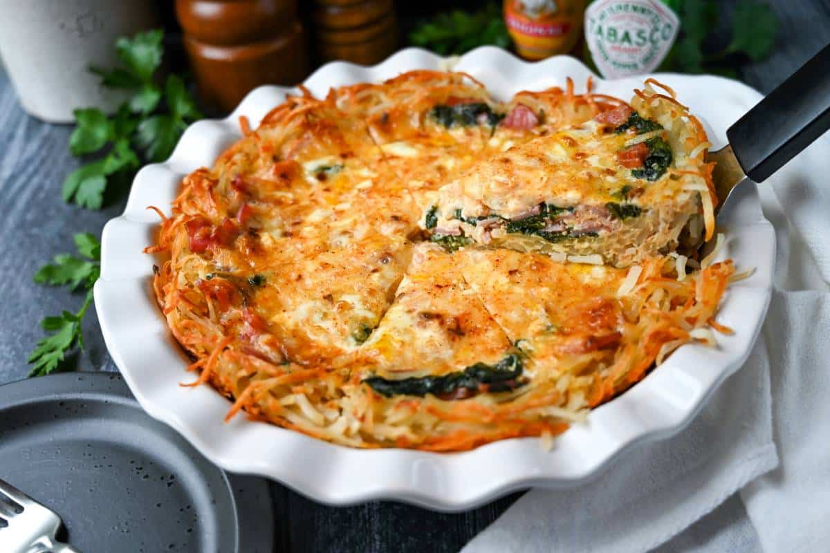 a slice of hashbrown quiche on a spatula with hot sauce behind it