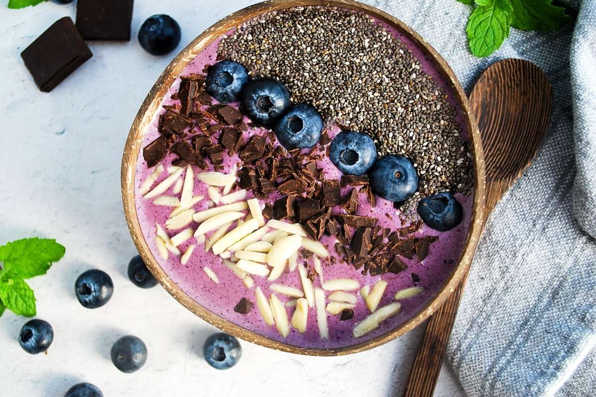 blueberry smoothie bowl with almonds, chocolate, chia seeds, and fresh blueberries in a coconut bowl with a spoon
