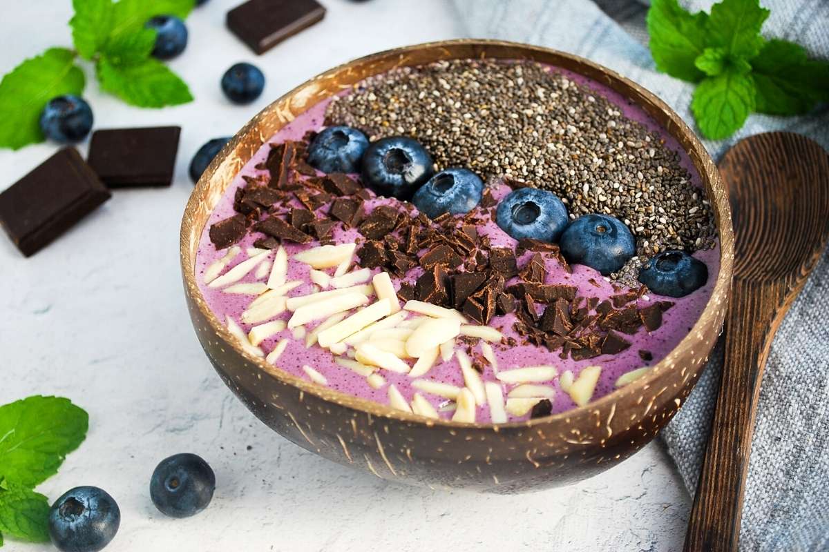 side view of a smoothie bowl topped with berries, chocolate, almonds, and chia seeds with mint, berries and chocolate in the background