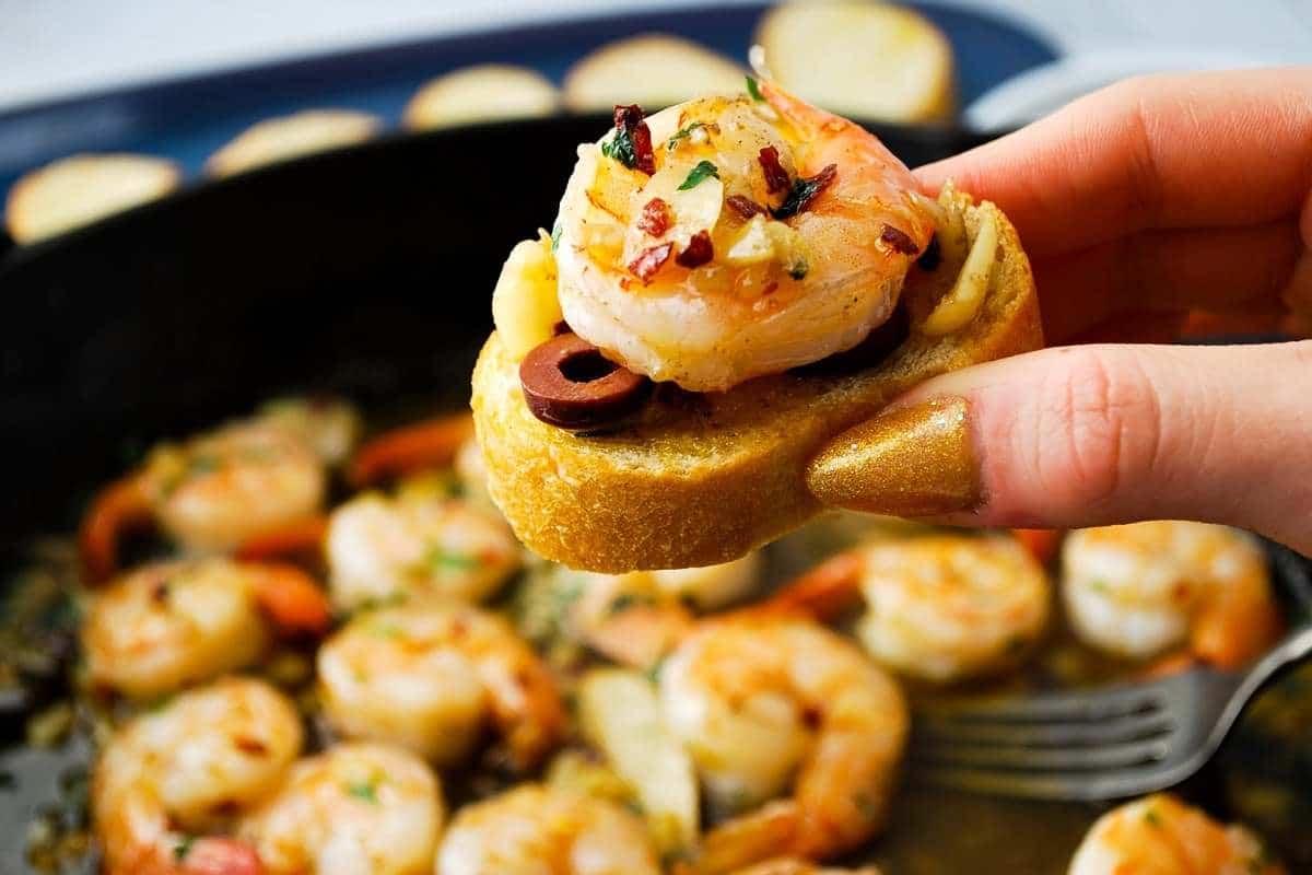 a prawn on top of a crusty baguette slice with olives, garlic, and olive oil