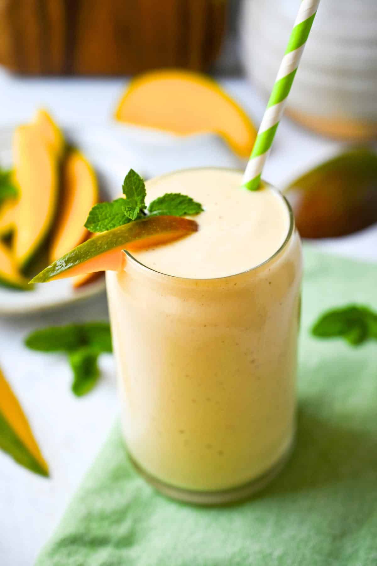a mango smoothie in a glass with a green striped straw with a cutting board and mango slices