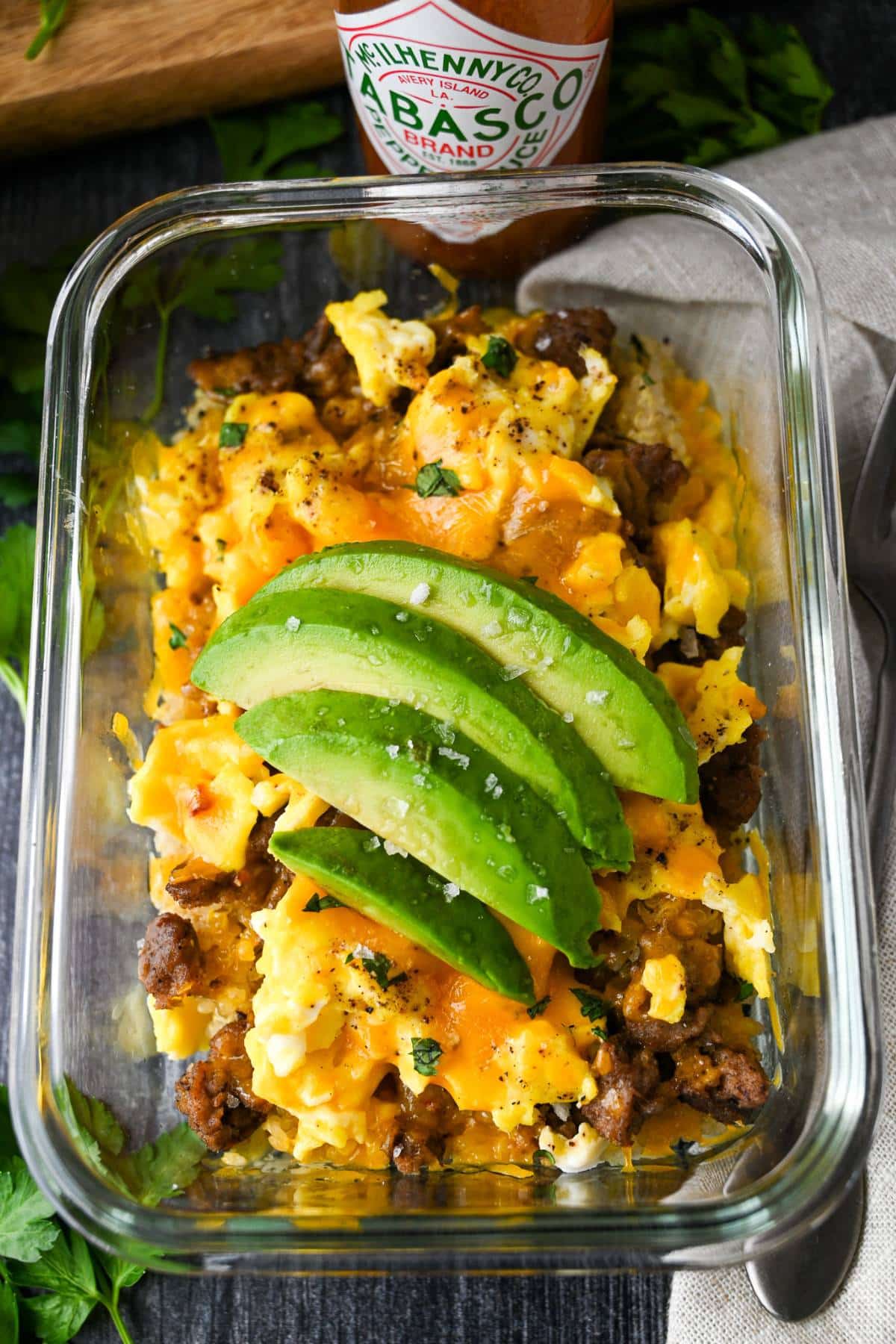 a breakfast bowl with eggs, cheese, turkey sausage and quinoa in a glass meal prep bowl with sliced avocado on top and a bottle of hot sauce behind it